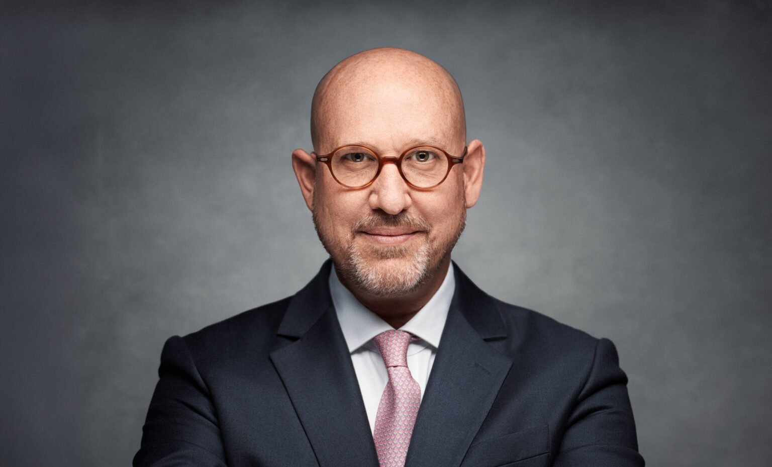 Bald man with round tortoiseshell glasses and grey stubble, navy suit, white shirt and pink patterned tie, smiling