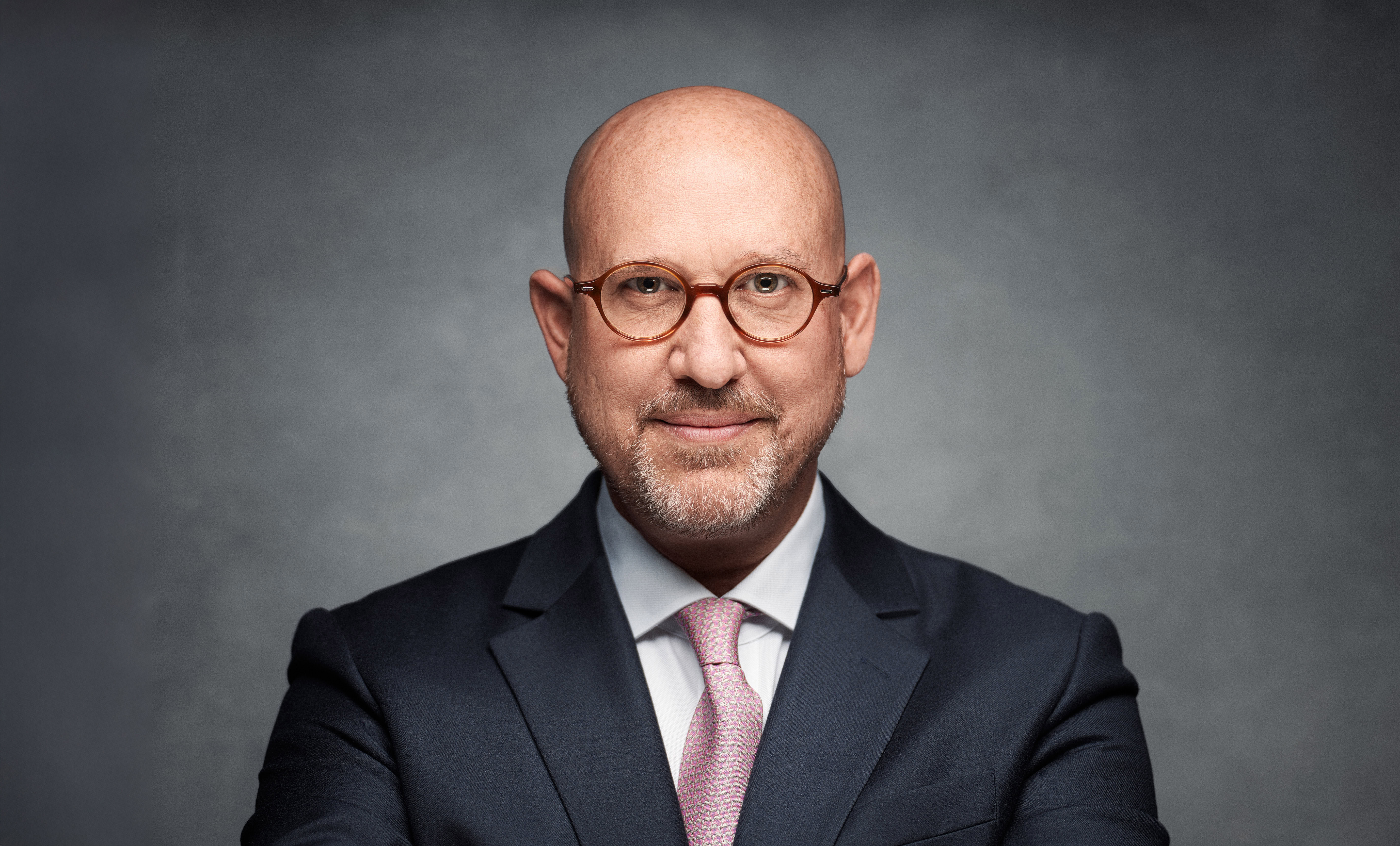 Bald man wearing glasses and a suit with a tie, looking directly at the camera.