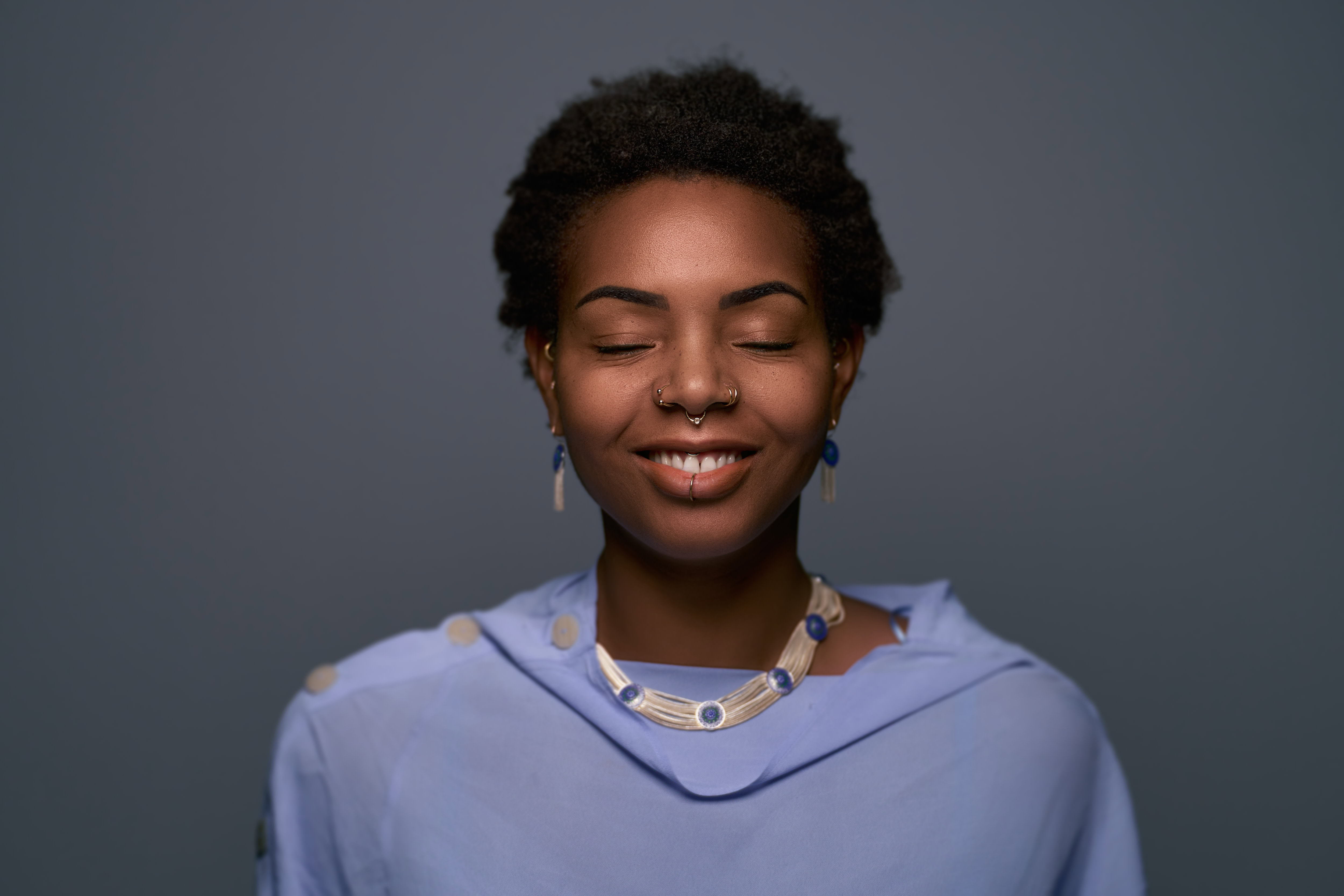 Corporate headshot featuring a smiling individual with closed eyes, wearing a light blue outfit and coordinating jewelry. Captured by Adam Toth of Tadam Photography, this image showcases professional studio lighting and a clean, minimalist background, creating a serene and approachable mood. Photographed in East London, this composition highlights natural expression and elegant style.