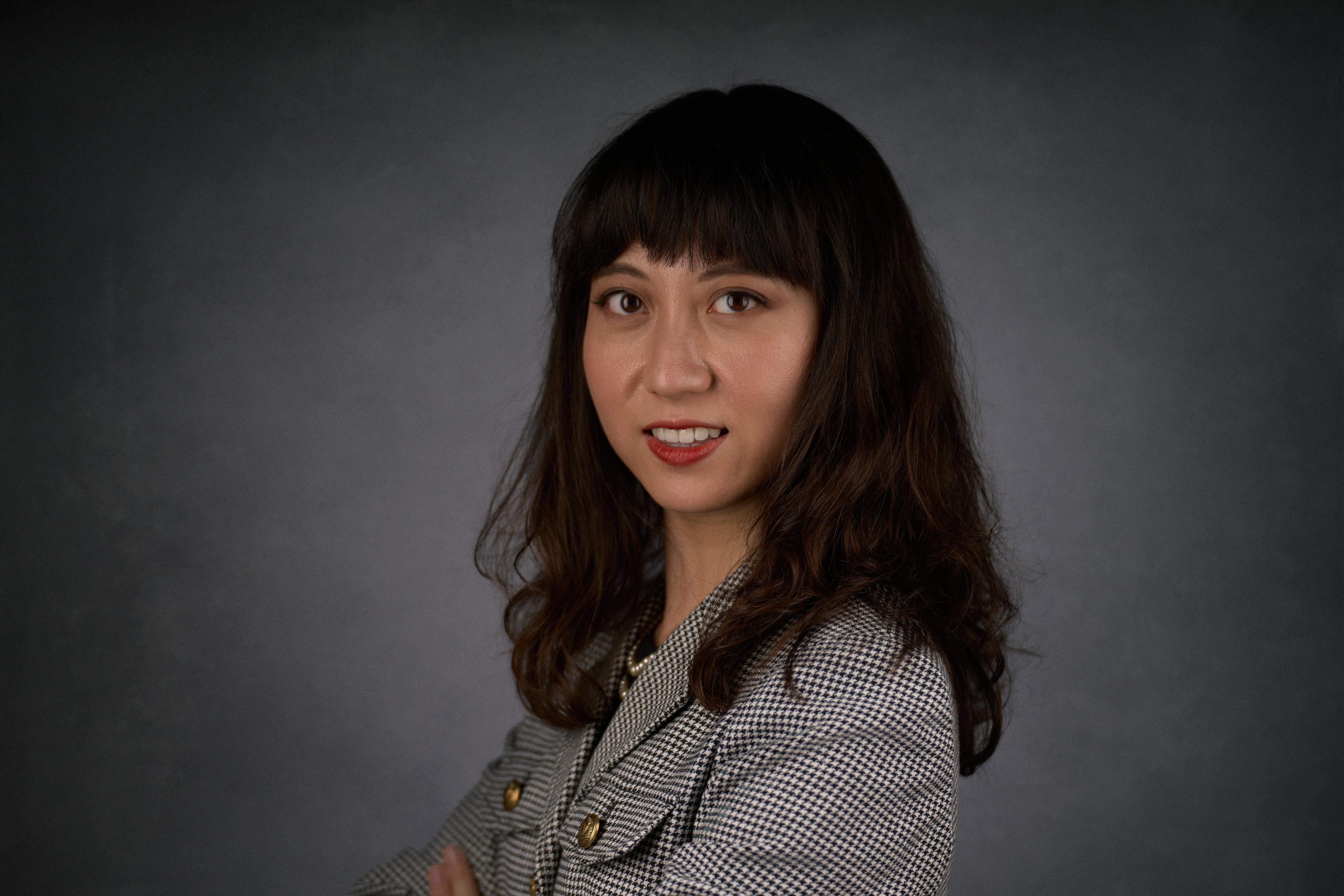 A woman with long hair wearing a checked blazer poses for a portrait against a dark background.