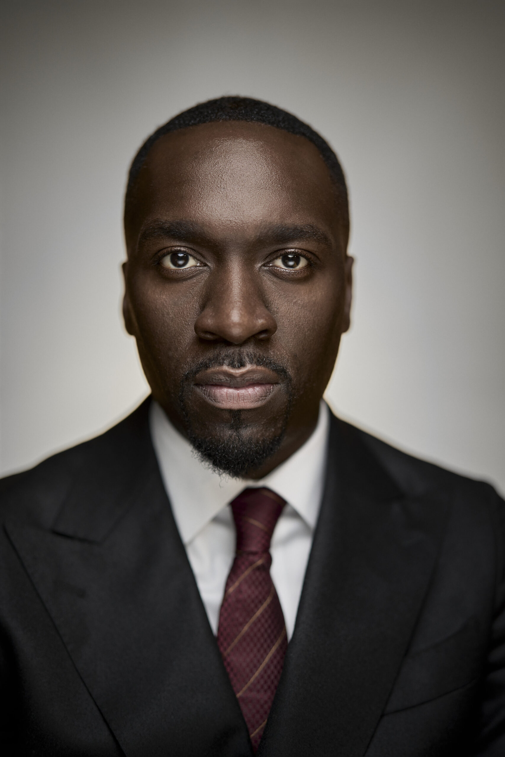 Corporate headshot featuring a confident professional in a tailored dark suit and maroon tie, captured with a neutral expression. The composition showcases crisp studio lighting against a clean background, embodying a polished and professional tone. Photographed by Adam Toth of Tadam Photography in London, highlighting expert portrait photography skills.