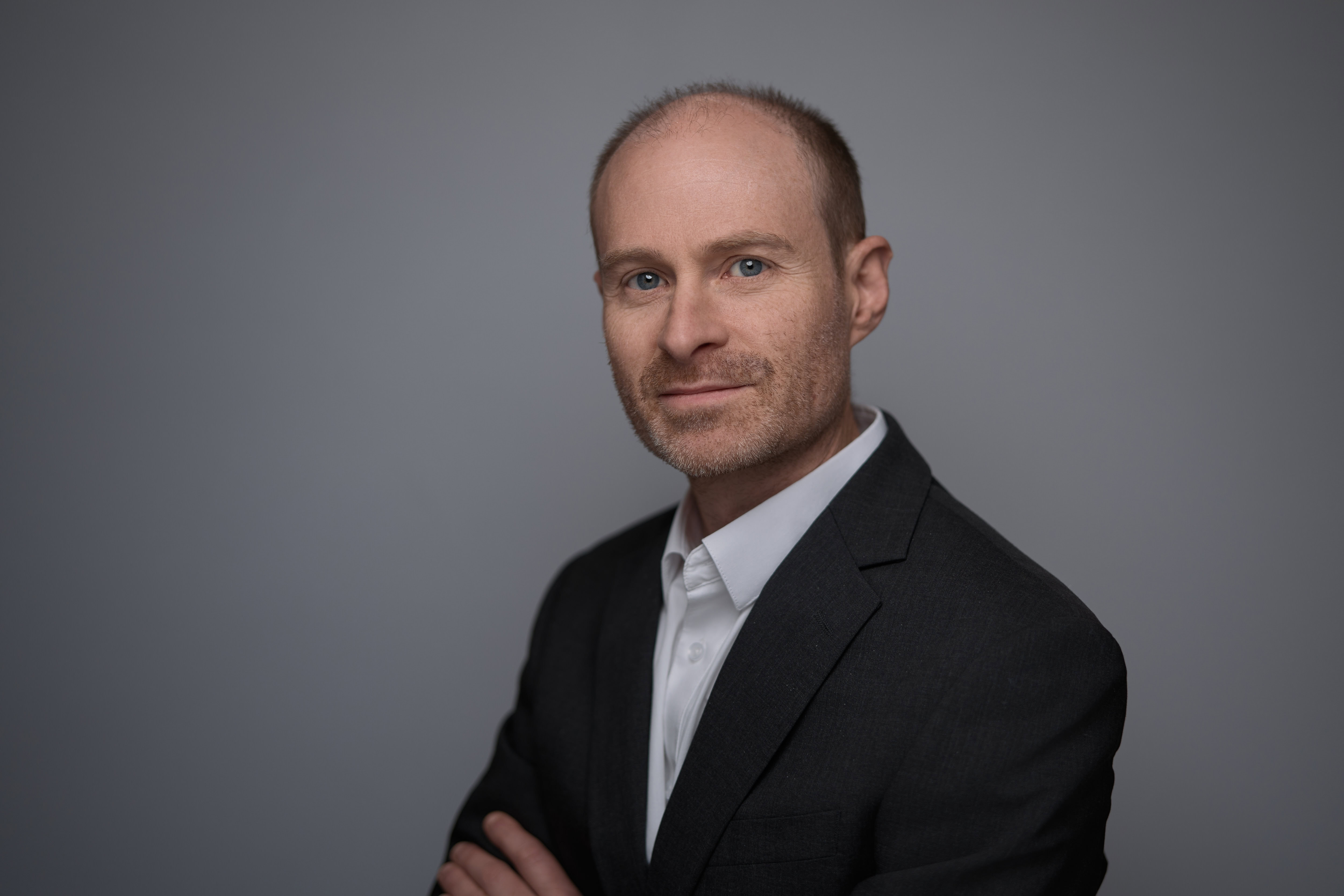 Corporate headshot of a man in a tailored suit, standing with arms crossed against a professional gray background. His confident expression is enhanced by studio lighting, creating a clean and polished look. Captured by Adam Toth of Tadam Photography in London, this portrait embodies professionalism and expertise, appealing to corporate clients in areas like East London and Canary Wharf.