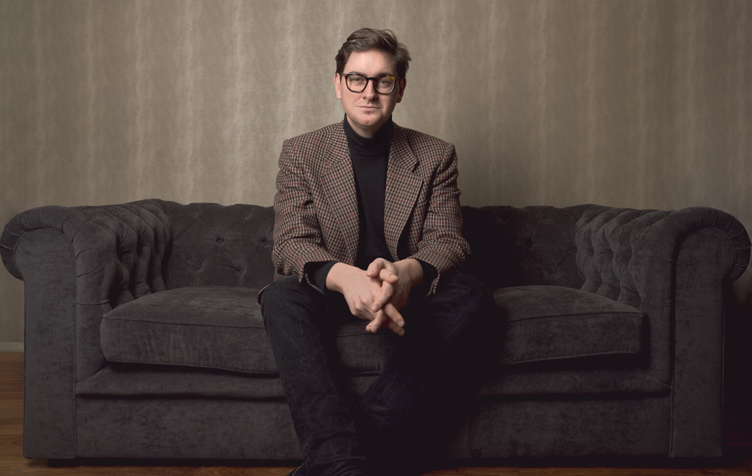 Johannes in a brown checked blazer and black rollneck, seated on a dark grey chesterfield sofa, hands clasped