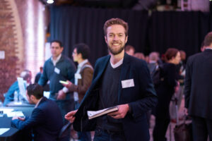 Man in dark blazer and grey sweater holding papers at an indoor event with people in background