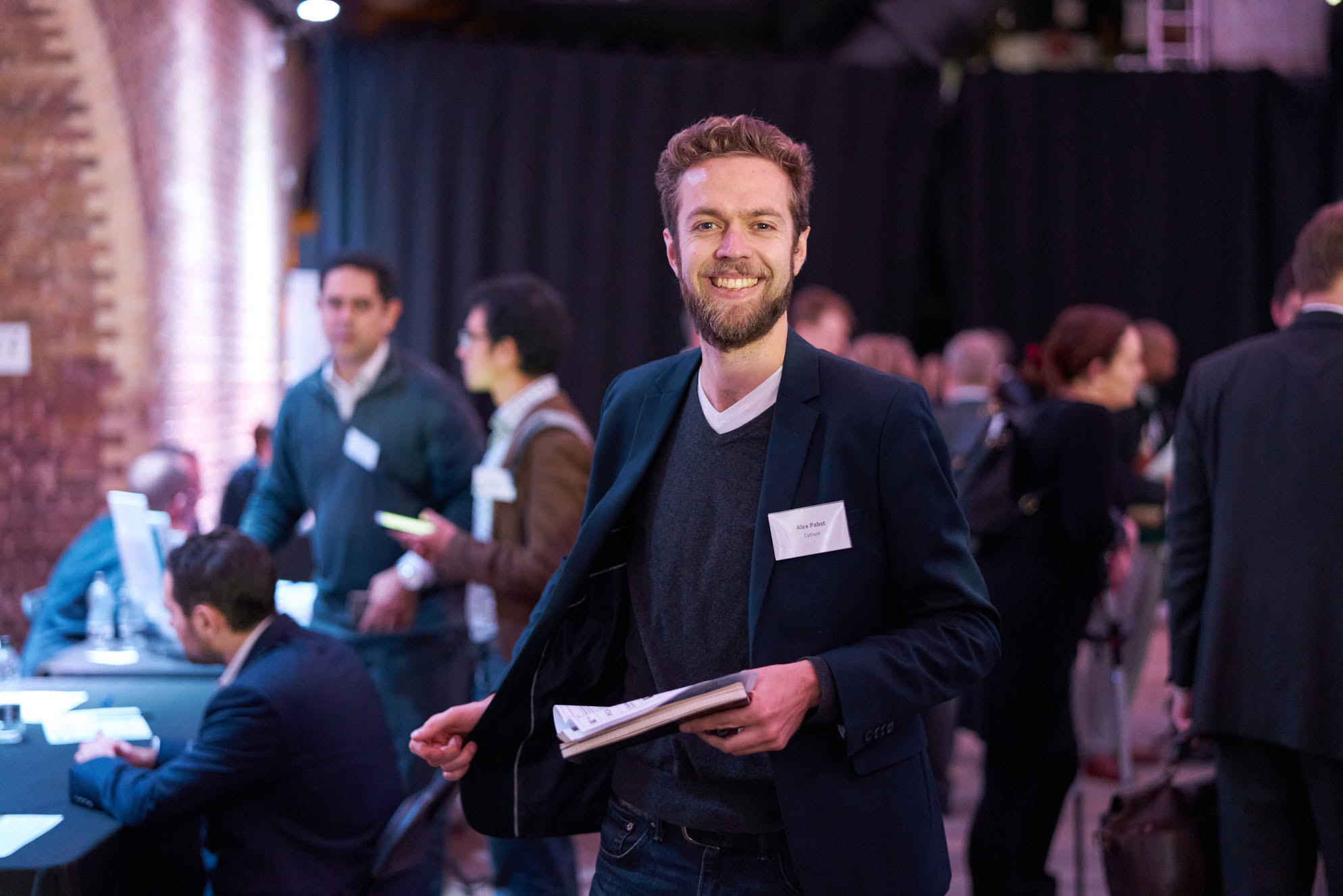 Man in dark blazer and grey sweater holding papers at an indoor event with people in background