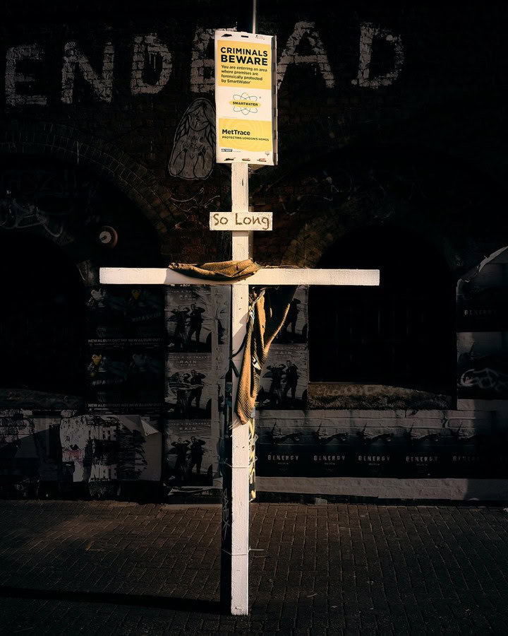 A white wooden cross with a yellow warning sign attached, located in an urban environment.
