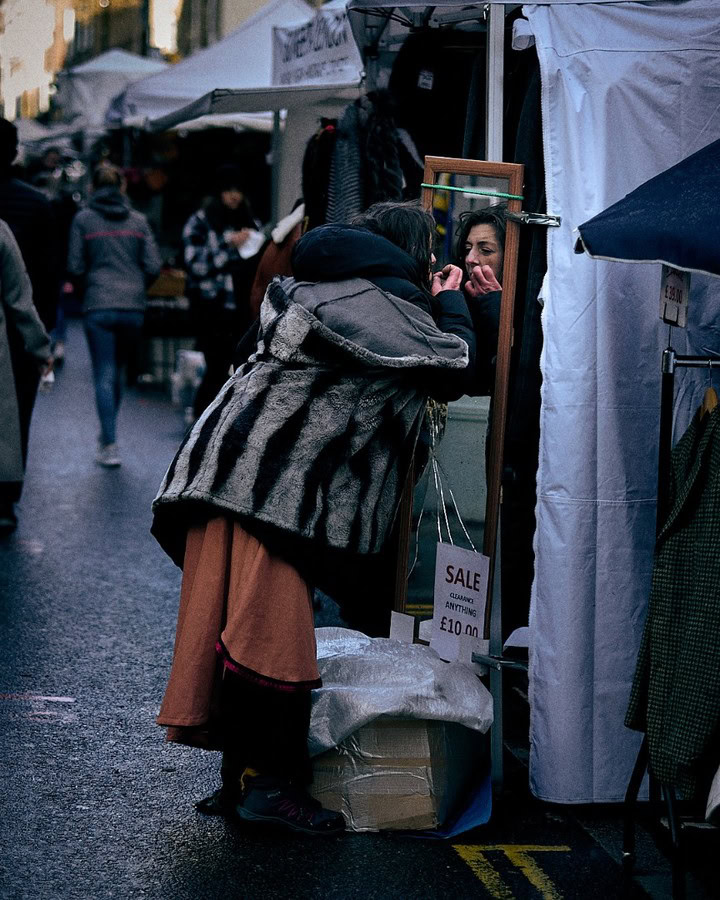 Street photography capture by Adam Toth of Tadam Photography in East London. A woman in layered clothing, including a striped coat, adjusts her makeup with a compact mirror at an outdoor market stall. Soft, natural lighting enhances the candid urban setting, capturing the lively atmosphere and authentic expressions of city life.