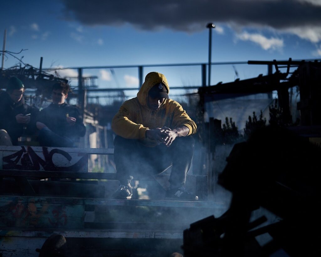 Individual wearing a yellow hoodie sitting on a wooden bench surrounded by smoke.