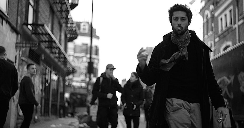 A man walking on a street holding a cigarette, with pedestrians in the background.