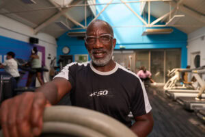 A vibrant gym portrait captured by Adam Toth of Tadam Photography in London, featuring an energetic individual in athletic gear. The subject is confidently engaging with exercise equipment under natural indoor lighting, enhancing the dynamic atmosphere. The modern gym setting with people in the background adds context to this East London location, emphasizing a lively fitness environment.