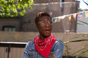 A candid portrait of a young individual wearing clear glasses and a vibrant red bandana set against an urban backdrop in London. Captured by Adam Toth of Tadam Photography, the subject stands confidently with an expressive pose, adorned in a blue denim jacket. The photo utilizes natural light filtering through surrounding greenery, creating a dynamic and engaging composition. The setting