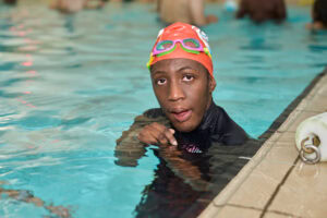 An enthusiastic swimmer in a vibrant orange swim cap and colorful goggles enjoys a moment in the pool, capturing the energy of youth sports photography. The clear water and subtle reflections convey a lively atmosphere. Shot by Adam Toth of Tadam Photography, this image skillfully utilizes natural lighting to highlight the subject’s determined expression. Captured in East London, the
