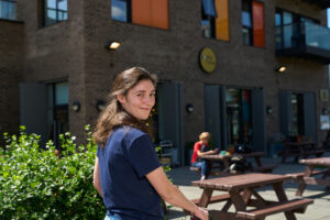 Outdoor portrait of a woman captured by Adam Toth of Tadam Photography in London. The subject stands with a relaxed, natural pose in a casual blue shirt, exuding a warm smile. Sunlight gently illuminates her face, enhancing her features against a vibrant green bush. The background reveals a modern brick building, contributing to the urban charm of an