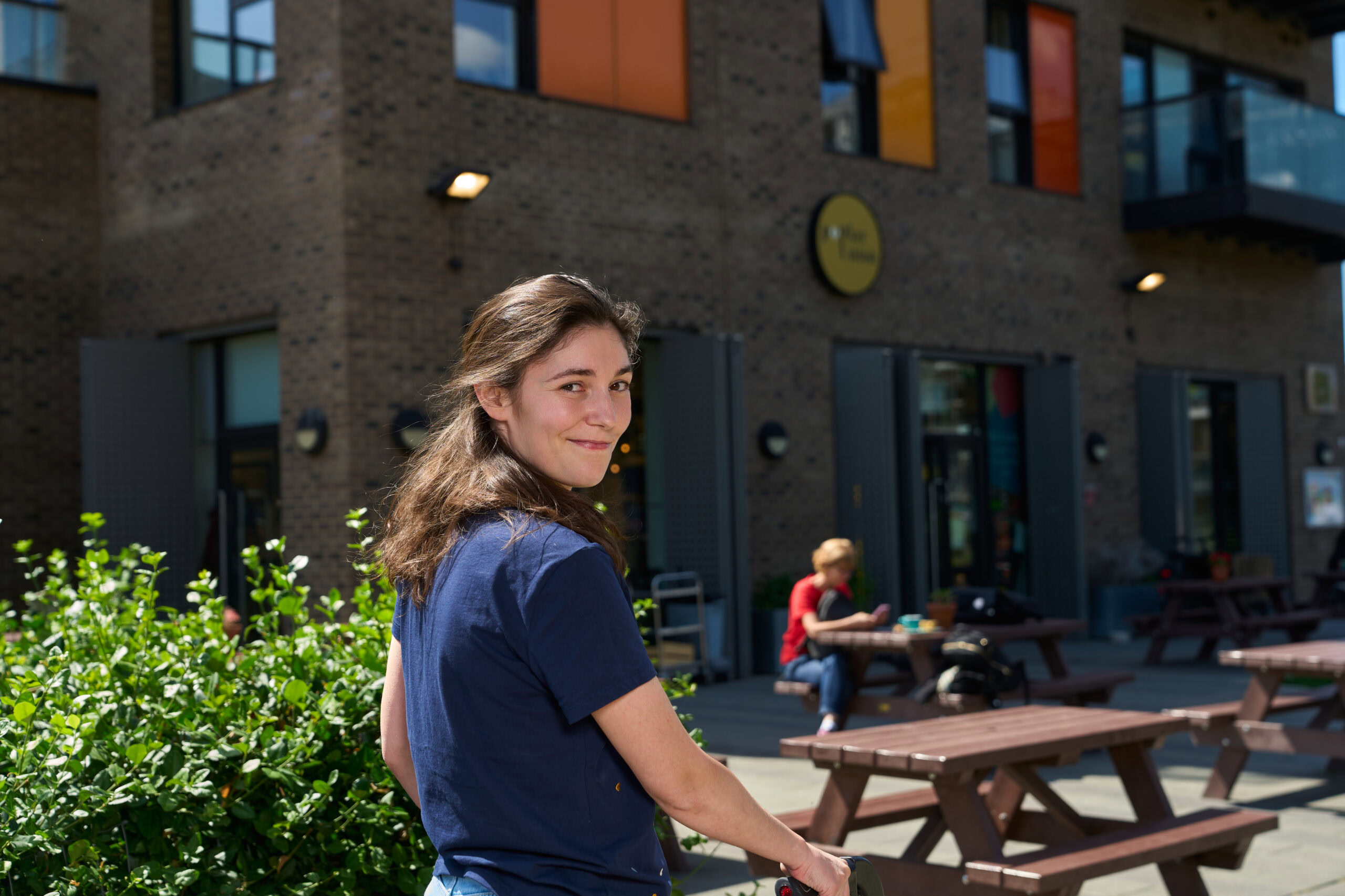 Outdoor portrait of a woman captured by Adam Toth of Tadam Photography in London. The subject stands with a relaxed, natural pose in a casual blue shirt, exuding a warm smile. Sunlight gently illuminates her face, enhancing her features against a vibrant green bush. The background reveals a modern brick building, contributing to the urban charm of an