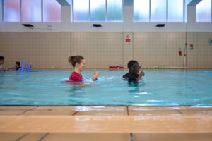 A young woman and a teenage boy engaged in swim instruction at an indoor pool in East London, captured by Adam Toth of Tadam Photography. The woman, confidently demonstrating a gesture, wears a vibrant pink swim shirt, while the boy, donning a swim cap and goggles, mirrors her actions. The scene is set against a backdrop of large,