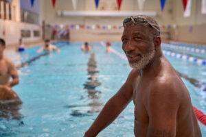An enthusiastic swimmer with a joyful expression stands by the edge of an indoor pool in London. The bright overhead lighting highlights his confident pose, while water droplets glisten on his skin. The background features blurred swimmers and vibrant pool flags. Captured by Adam Toth of Tadam Photography, this image conveys a lively and energetic atmosphere.