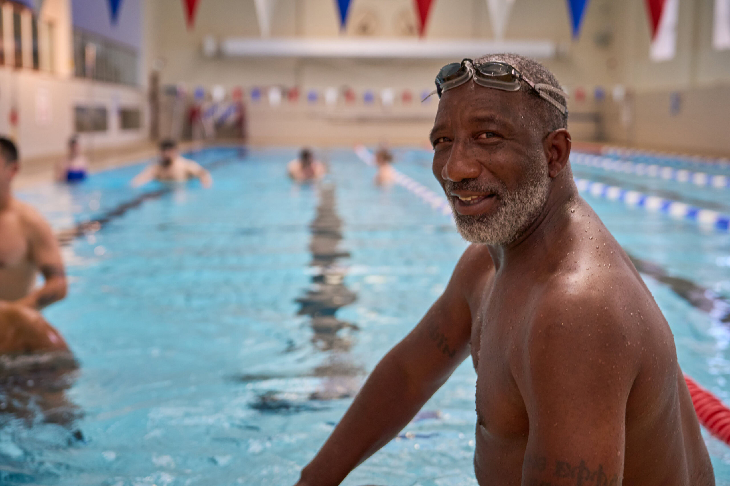 An enthusiastic swimmer with a joyful expression stands by the edge of an indoor pool in London. The bright overhead lighting highlights his confident pose, while water droplets glisten on his skin. The background features blurred swimmers and vibrant pool flags. Captured by Adam Toth of Tadam Photography, this image conveys a lively and energetic atmosphere.