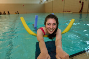 Corporate headshot of a smiling woman swimming with a yellow pool noodle in a London indoor pool captured by Adam Toth at Tadam Photography. The subject's joyful expression and natural pose create an inviting atmosphere. The image features soft, natural lighting reflecting off the water, adding a lively and dynamic mood to the composition. Ideal for portraying a relaxed and approachable