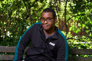 Corporate headshot captured by Adam Toth of Tadam Photography in London. The subject, dressed in a casual black and teal zip-up jacket, sits confidently on a wooden bench amid lush greenery. Natural light creates an inviting and approachable mood, with vibrant foliage providing a contrasting backdrop. Ideal for personal branding or professional networking profiles.