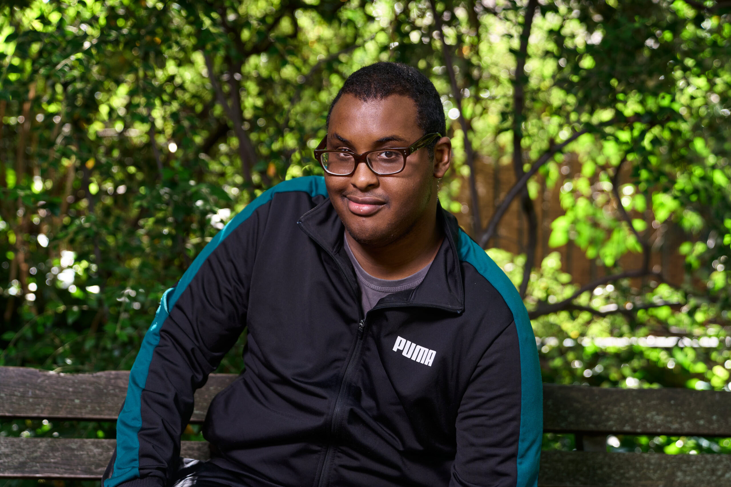 Corporate headshot captured by Adam Toth of Tadam Photography in London. The subject, dressed in a casual black and teal zip-up jacket, sits confidently on a wooden bench amid lush greenery. Natural light creates an inviting and approachable mood, with vibrant foliage providing a contrasting backdrop. Ideal for personal branding or professional networking profiles.