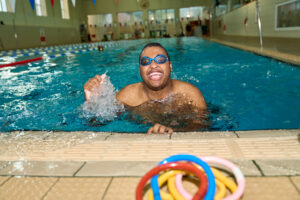 Swimmer enjoying a moment in a London pool, captured by Adam Toth of Tadam Photography. The subject wears goggles and expresses joy, with water splashing around. The bright indoor setting and natural lighting enhance the vibrant atmosphere, reflecting the enthusiasm of a recreational swim session in East London.