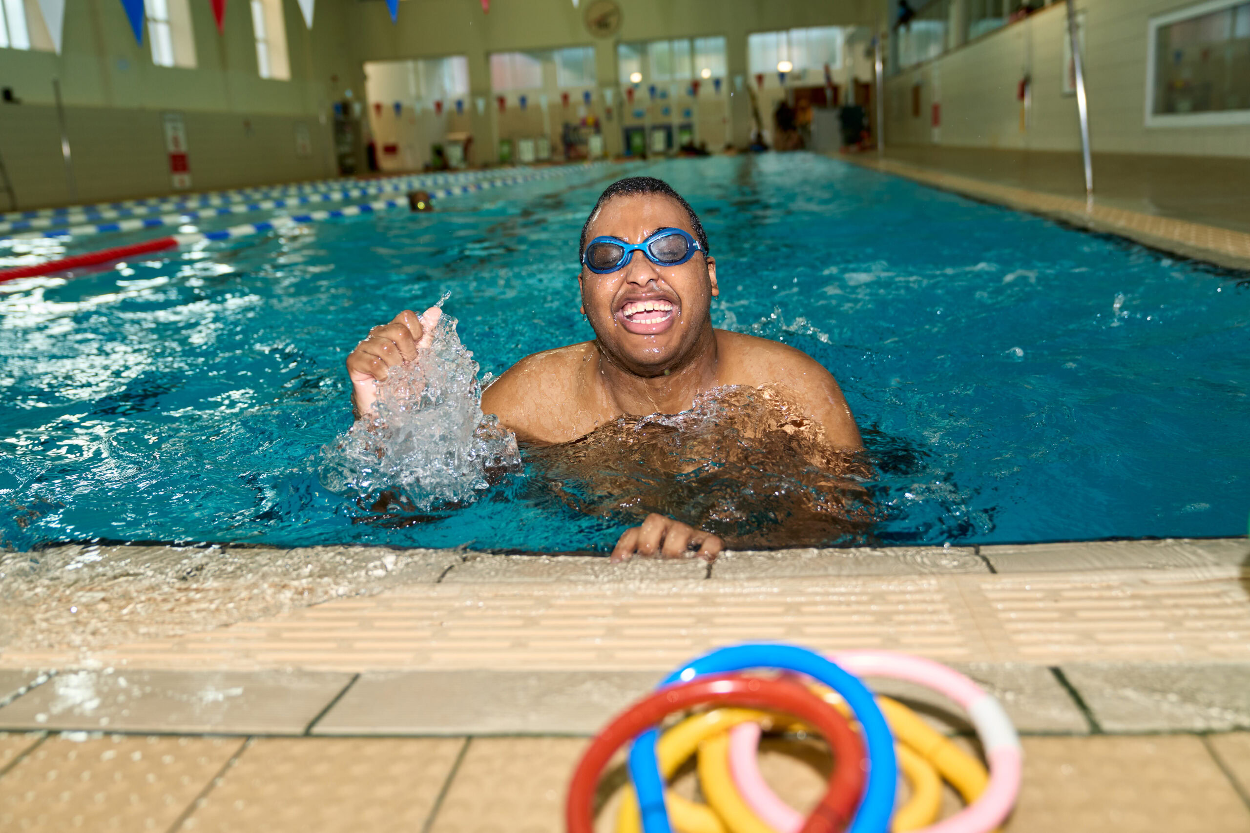 Swimmer enjoying a moment in a London pool, captured by Adam Toth of Tadam Photography. The subject wears goggles and expresses joy, with water splashing around. The bright indoor setting and natural lighting enhance the vibrant atmosphere, reflecting the enthusiasm of a recreational swim session in East London.