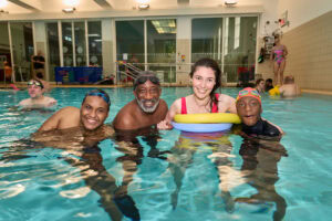 Group swimming lesson photo captured by Adam Toth of Tadam Photography, featuring four cheerful adults in a swimming pool in London. The subjects display joyful expressions and wear swim gear, illuminated by bright indoor lighting. This dynamic and engaging shot highlights the inclusive and supportive environment of the session. The modern leisure center setting adds to the vibrant mood, typical of Adam