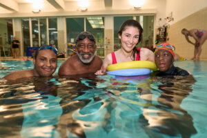 Family enjoying a swim session, captured by Adam Toth at Tadam Photography in London. The diverse group smiles warmly in the pool, exuding joy and connection during this candid family photography session. The vibrant indoor lighting accentuates the inviting atmosphere, with reflections shimmering on the water. This engaging scene, set in East London, highlights the harmonious blend