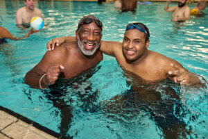 A joyful and candid moment captured by Adam Toth of Tadam Photography, highlighting an engaging family gathering in a London swimming pool. The scene centers on two smiling individuals in vibrant swimwear, embodying camaraderie and lightheartedness. The water creates dynamic reflections and adds a playful mood to the image. Professional lighting enhances the lively expressions and cheerful atmosphere