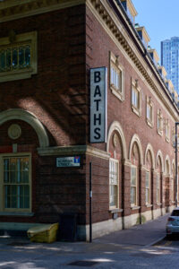 Alt text: A captivating architectural photograph by Adam Toth of Tadam Photography, featuring a historic brick building on Norman Street in East London. The composition highlights the building's intricate arches and ornate windows under soft, natural lighting. The image captures the urban charm of the area, with a clear blue sky and distant modern skyscrapers adding a contrasting backdrop.