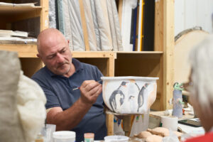 A man in a blue polo shirt painting a black and white pattern onto a ceramic pot, in a workshop with wooden shelving