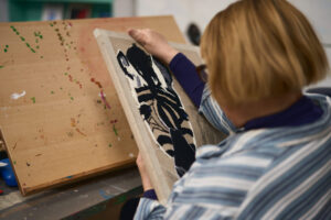 Woman working on a black-and-white print at a paint-spattered board, seen over her shoulder