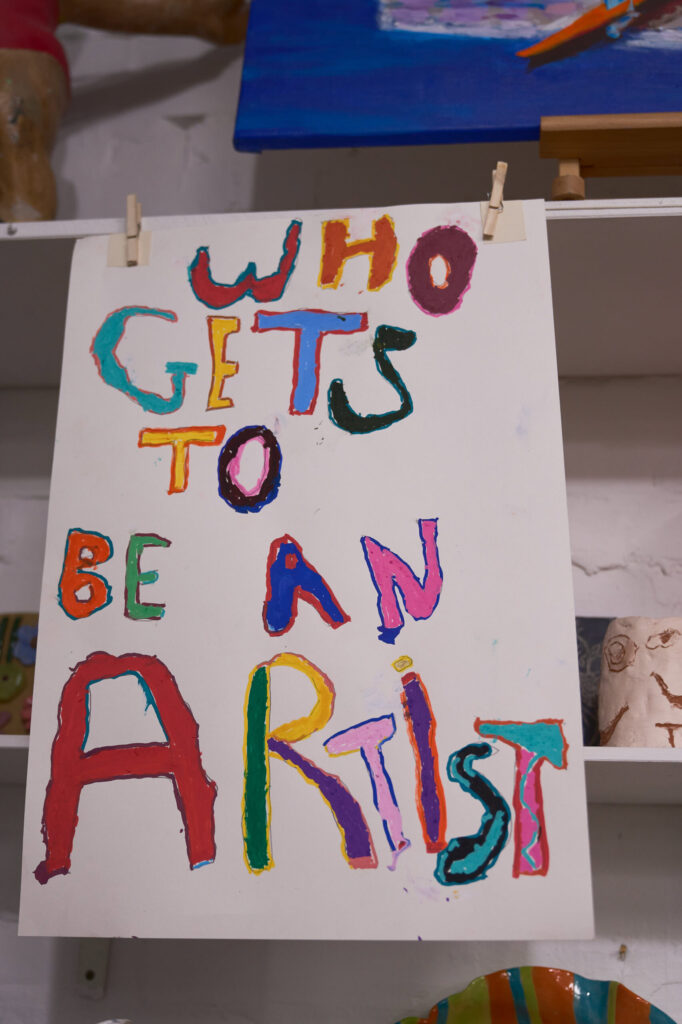 Hand-painted sign reading Who Gets To Be An Artist pegged above an art studio shelf