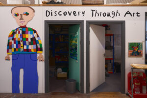 Painted life-size figure in a patchwork jumper beside Discovery Through Art lettering in a studio