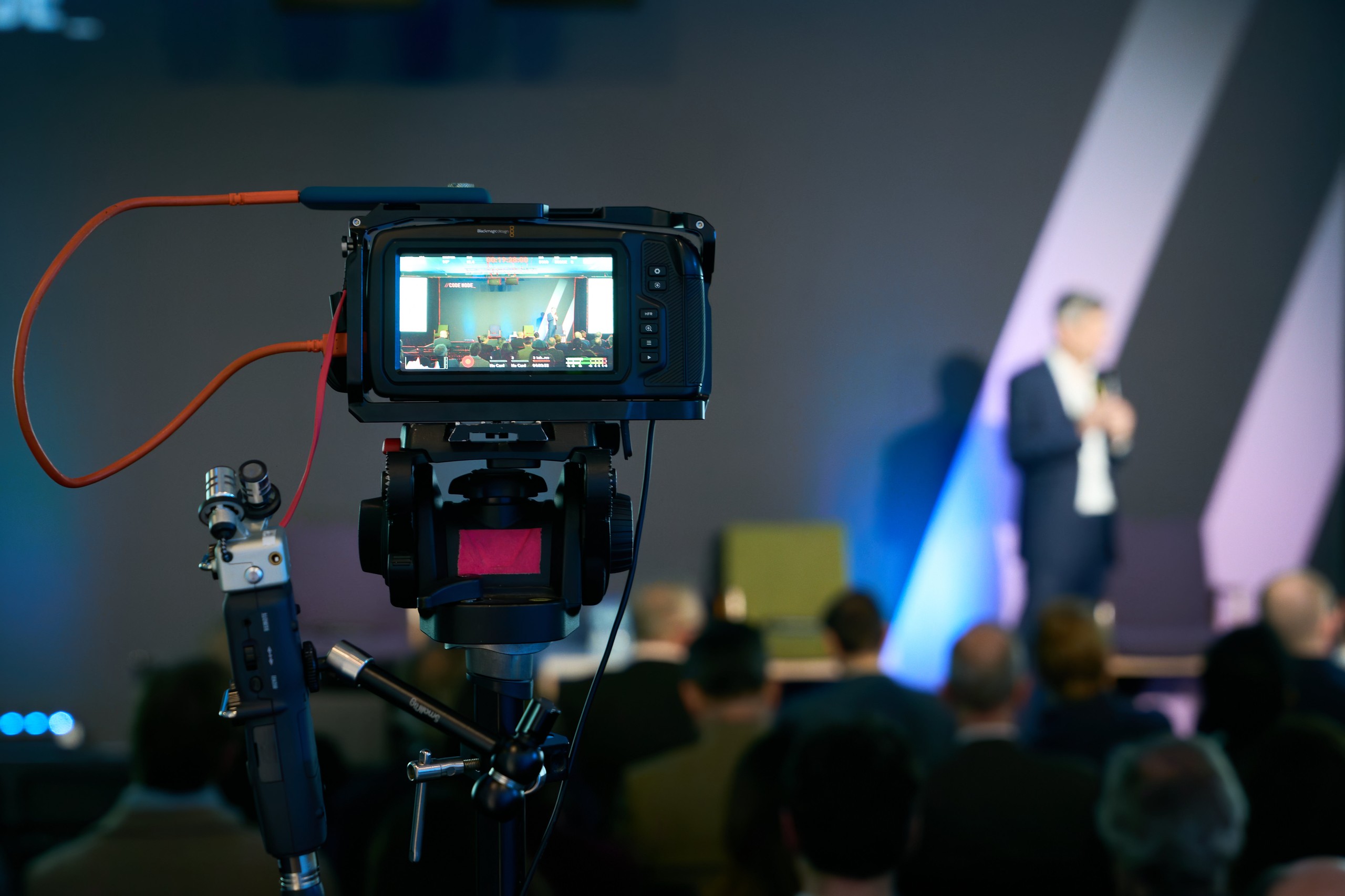 Video camera on a tripod filming a speaker on stage, its screen showing the stage, the audience in silhouette