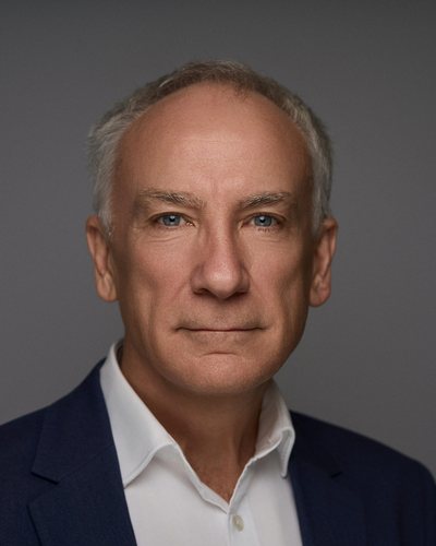 Headshot of a man with short white hair in a navy blazer and white shirt, steady look on a grey backdrop