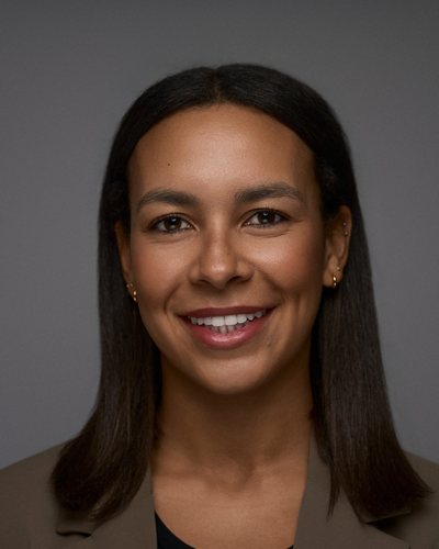 Headshot of a woman with long straight dark hair in a taupe blazer over a black top, smiling, grey backdrop