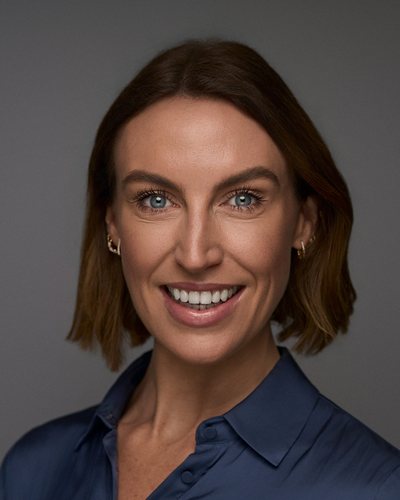 Headshot of a woman with a short brown bob in a navy satin shirt, broad smile on a grey backdrop