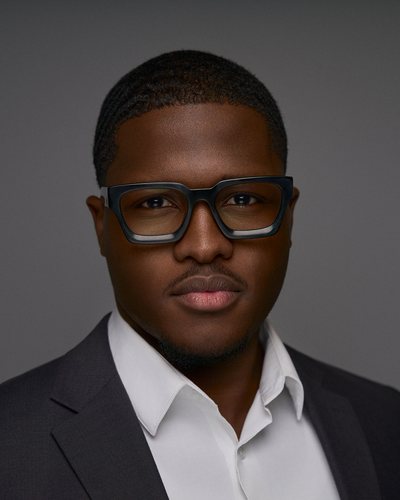Headshot of a man with bold black glasses in a charcoal blazer and white open-collar shirt, grey backdrop