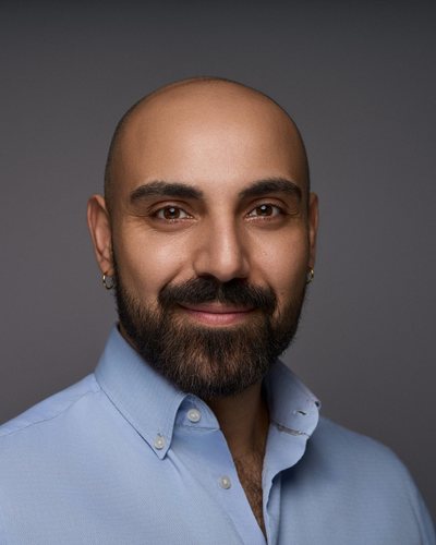 Headshot of a man with a shaved head and dark beard in a light blue open-collar shirt, grey backdrop