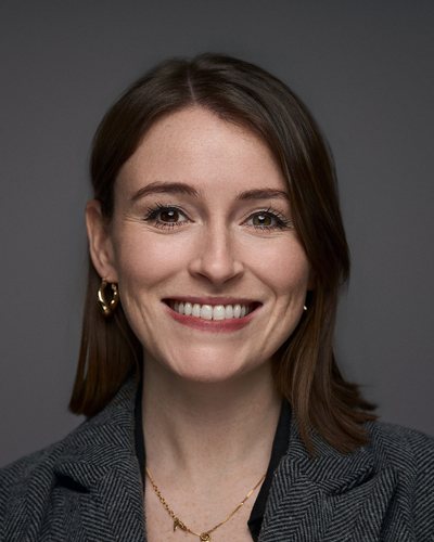 Headshot of a woman with shoulder-length brown hair in a grey herringbone blazer, broad smile on a grey backdrop