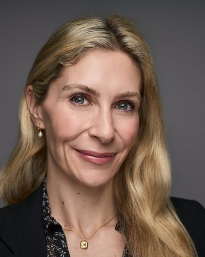 Headshot of a woman with long wavy blonde hair in a black blazer and patterned blouse, smiling on a grey backdrop