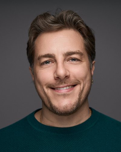 Headshot of a man with swept-back hair and stubble in a dark green jumper, broad smile on a grey backdrop