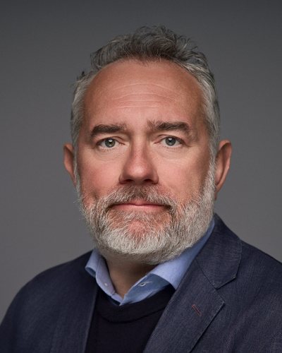 Headshot of a man with grey hair and a white beard in a navy jacket over a jumper and shirt, grey backdrop