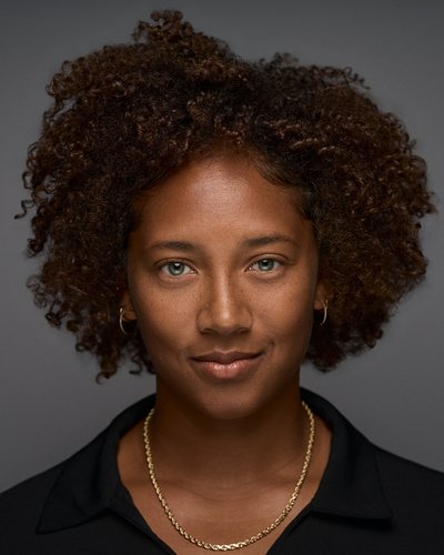 Headshot of a woman with curly natural hair in a black shirt and gold chain, slight smile on a grey backdrop