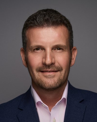 Headshot of a man with short dark hair in a navy blazer over a pale pink shirt, grey backdrop